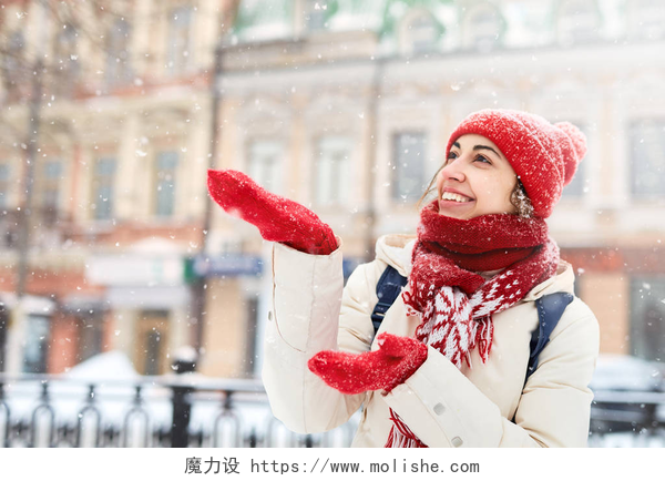 在寒冷的冬天一个穿着全副武装的女人在室外看着雪花落下穿着白色羽绒服、红色帽子、围巾和手套的快乐微笑的妇女走在雪地上，在城市暴风雪过后捕捉雪花。专注于手套